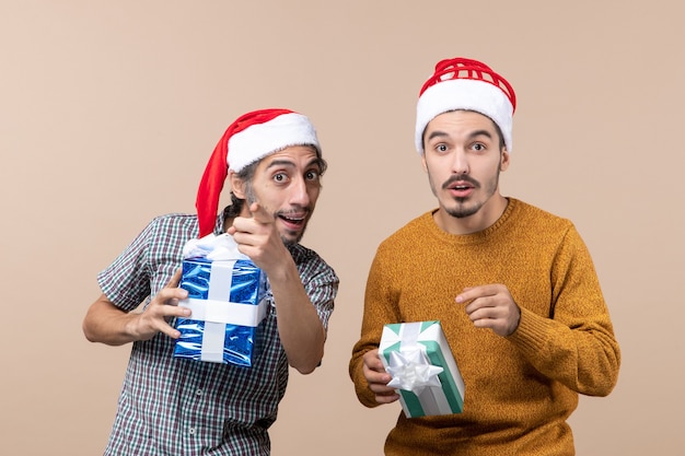 Вид спереди два заинтересованных парня указывают пальцем, показывая что-то и держат рождественские подарки на бежевом изолированном фоне