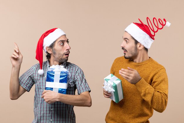 Front view two interested guys showing something to each other holding christmas presents on beige isolated background