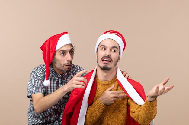 Front view two interested guys with santa hats looking at something on beige isolated background