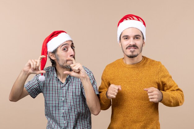 Вид спереди двух заинтересованных парней в новогодних шапках, один делает знак камеры руками на бежевом изолированном фоне