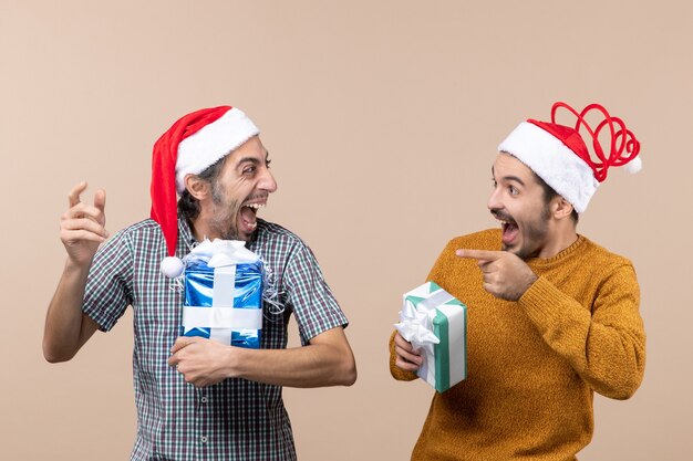Front view two laughing guys with santa hats holding christmas presents on beige isolated background