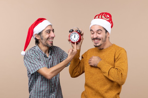 Front view two laughing men one holding an alarm clock and the other showing time on beige isolated background