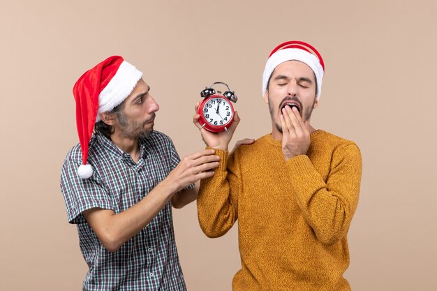 Front view two men one holding an alarm clock while yawning on beige isolated background