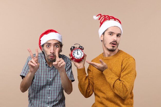 Front view two men one showing time with his fingers and the other holding an alarm clock on beige isolated background