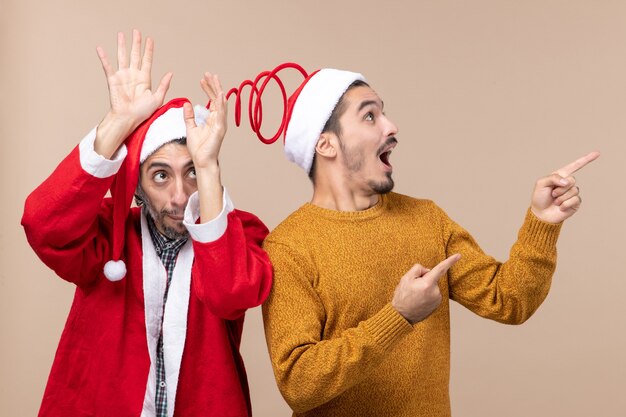 Front view two men with santa hats one raising his hands to his head and the other showing direction on beige isolated background