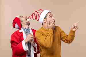 Free photo front view two men with santa hats one trying to catch hats pompom and the other showing direction on beige isolated background