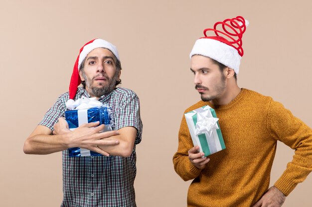 Front view two prying guys wearing santa hats and holding their presents on beige isolated background