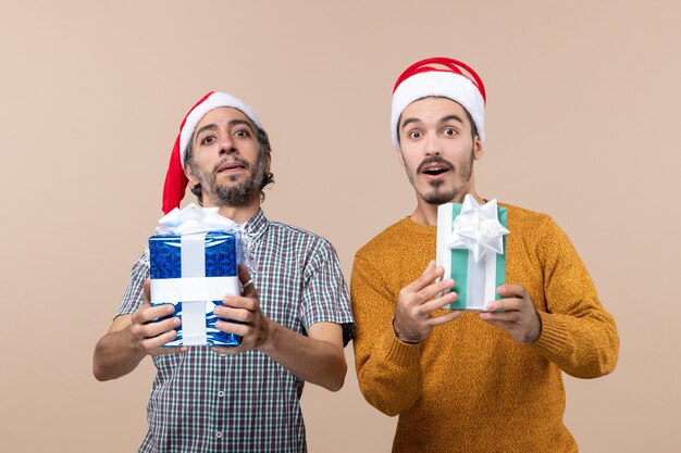 Front view two satisfied guys with santa hats and holding christmas presents on beige isolated background