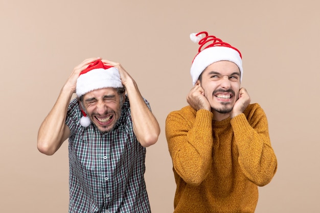 Front view two scared guys with santa hats one putting his hands on his head the other on his ears standing on beige isolated background