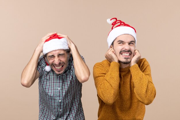 Front view two scared guys with santa hats one putting his hands on his head the other on his ears standing on beige isolated background