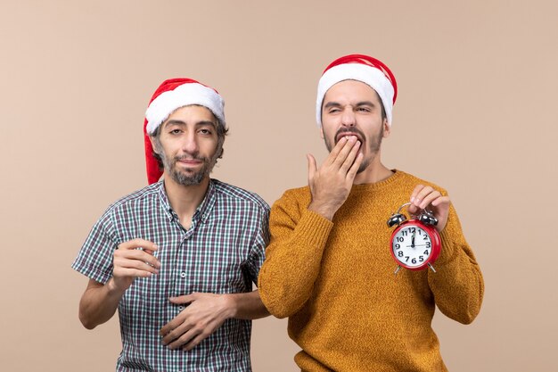 Front view two sleepy men one putting hand to his chest and the other yawning on beige isolated background