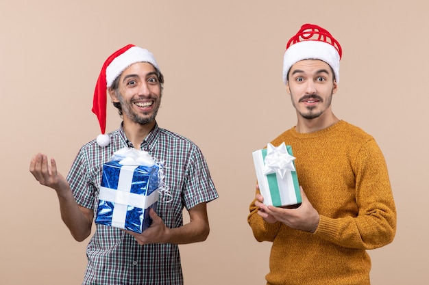 Front view two smiled guys wearing santa hats and holding christmas presents on beige isolated background