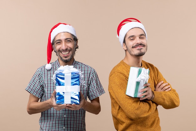 Front view two smiling guys wearing santa hats and holding christmas presents