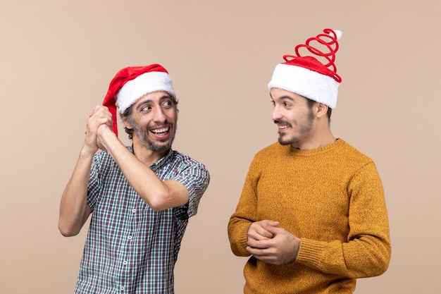 Front view two smiling guys with santa hats looking at each other on beige isolated background