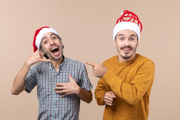 Front view two smiling guys with santa hats one making call me phone sign and the other point finger showing him on beige isolated background