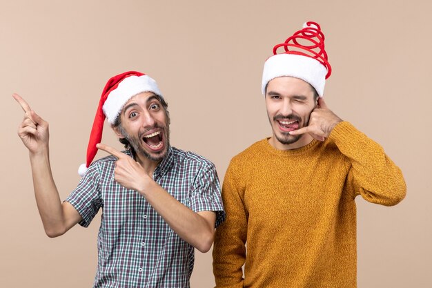 Front view two smiling guys with santa hats one showing something and the other making call me phone sign on beige isolated background