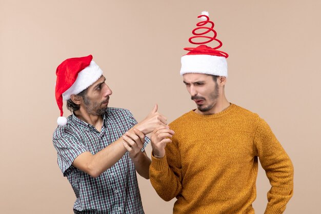 베이지 색 격리 된 배경에 다른 가입 엄지 손가락을 보여주는 산타 모자와 함께 전면보기 두 이상한 남자