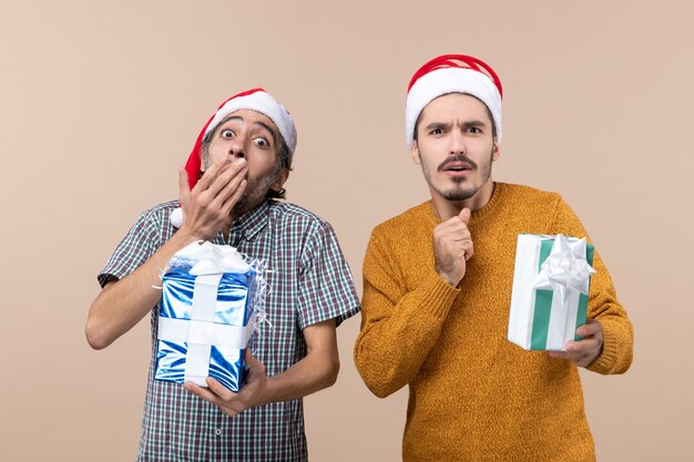 Front view two wondered guys with santa hats and holding presents one putting hand on his mouth on beige isolated background