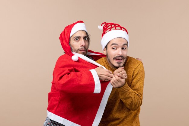 Вид спереди двух рождественских парней в пальто санта-клауса, греющих своего друга на бежевом изолированном фоне