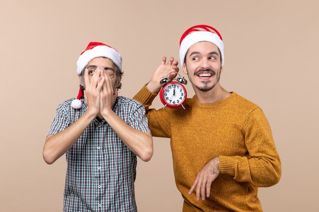 Вид спереди двух рождественских мужчин, один прикрывающих один глаз руками, а другой с будильником на бежевом изолированном фоне