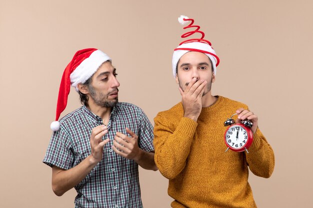 Вид спереди двух рождественских мужчин, один из которых держит будильник и прикрывает рот рукой на бежевом изолированном фоне
