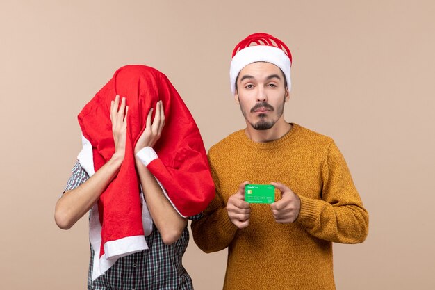 Front view two xmas men one holding credit card and the other covering his head with santa coat on beige isolated background