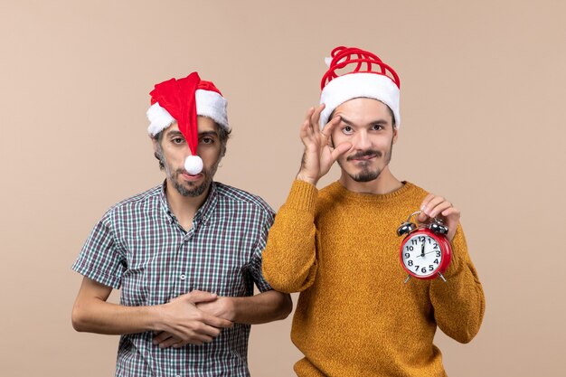 Front view two xmas men one putting hands on his chest the other holding an alarm clock on beige isolated background