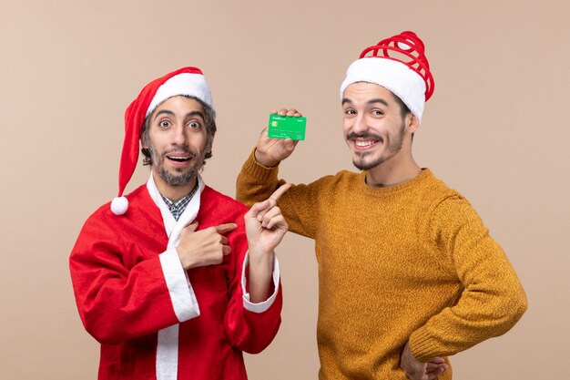 正面図2人のクリスマスの男性1人はサンタのコートを着ており、もう1人はベージュの孤立した背景に腰に手を置いてクレジットを持っています