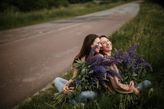 Вид спереди двух молодых брюнеток, сидящих на траве с большими свежими букетами диких люпинов возле дороги и искренне улыбающихся