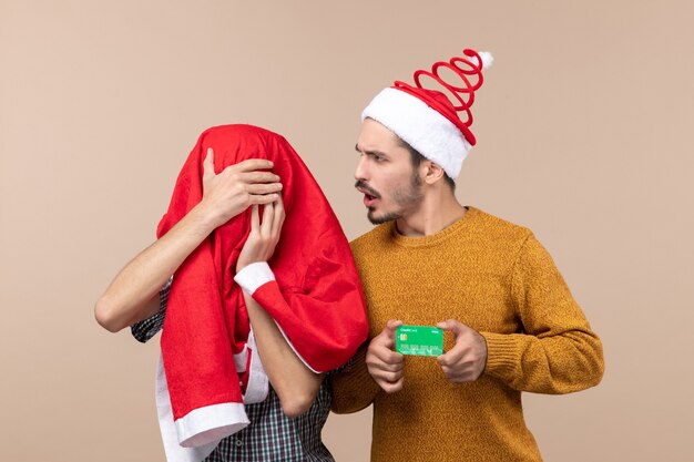 Front view two young men one holding credit card and the other covering his head with santa coat on beige isolated background