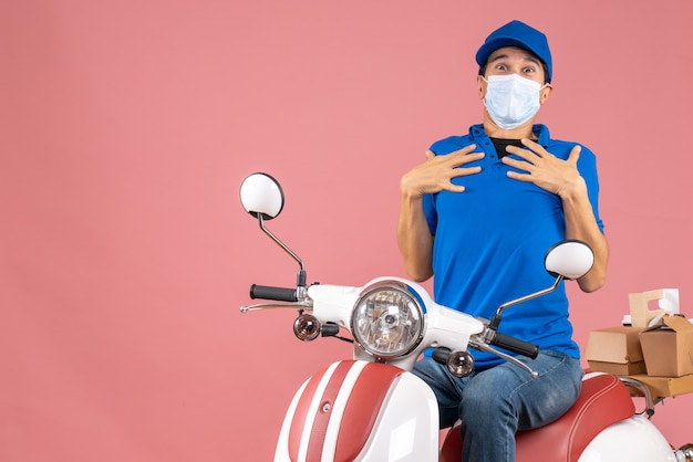 Front view of uncertain courier man in medical mask wearing hat sitting on scooter delivering orders on pastel peach background