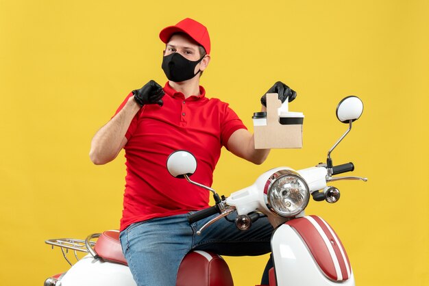 Front view of uncertain courier man wearing red blouse and hat gloves in medical mask sitting on scooter showing orders pointing himself