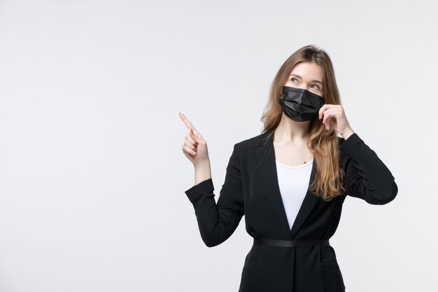 Front view of uncertain unsure young lady in suit wearing surgical mask and pointing up on white