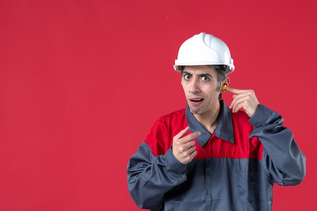 Vista frontale di un giovane lavoratore insoddisfatto in uniforme con elmetto e con tappi per le orecchie sul muro rosso isolato