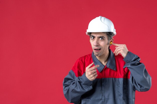 Front view of unsatisfied young worker in uniform with hard hat and wearing earplugs on isolated red wall