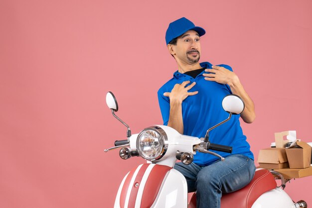 Front view of unsure delivery guy wearing hat sitting on scooter pointing himself on pastel peach background