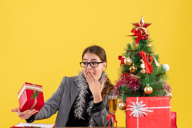 素晴らしい驚きのクリスマスツリーとギフトカクテルでギフトを見てテーブルに座っている眼鏡を持ってさまよう少女の正面図