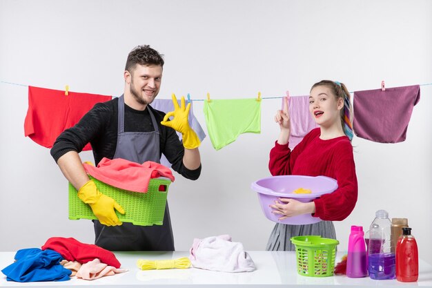 オーケーのサインを作る洗濯時間の男性とテーブルの洗濯かごの後ろに立ってテーブルの上で物を洗う妻の正面図
