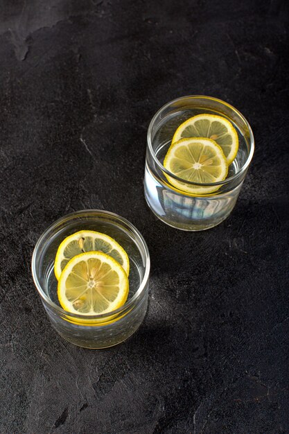 A front view water with lemon drink with sliced lemons inside transparent glasses on the dark