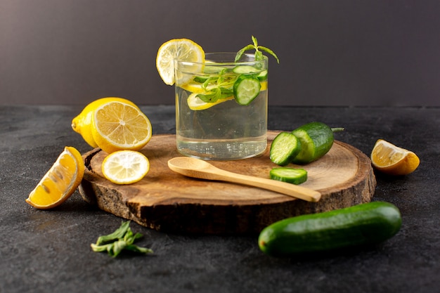 A front view water with lemon fresh cool drink inside glass with green leaves with ice cubes sliced lemons cucumber on the dark
