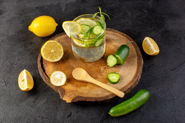 A front view water with lemon fresh cool drink inside glass with green leaves with ice cubes with sliced lemons cucumber on the dark