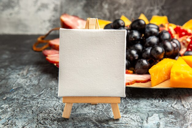 Front view white canvas on wooden easel grapes meat slices on wood plate on dark