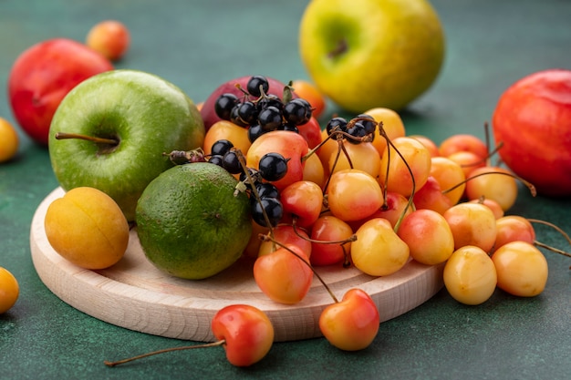 Front view of white cherry with black currant lime peach and a green apple on a stand on a green surface