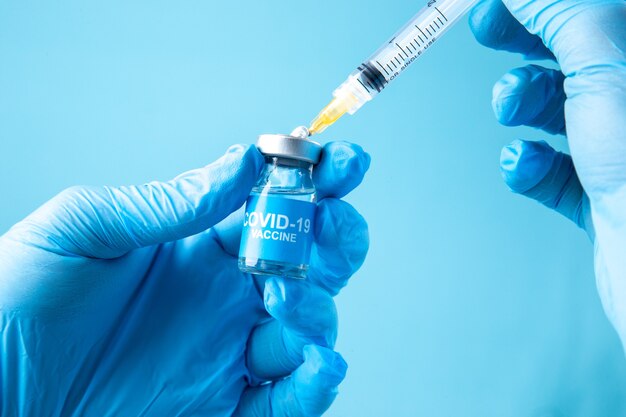 파란색 물결 배경에 covid-백신이 있는 닫힌 앰플과 주사기를 들고 있는 흰색 장갑의 전면 보기
