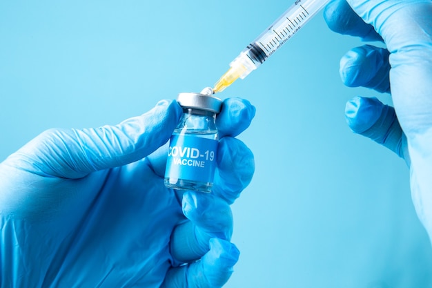 青い波の背景にcovid-ワクチンと閉じたアンプルと注射器を持っている手を身に着けている白い手袋の正面図