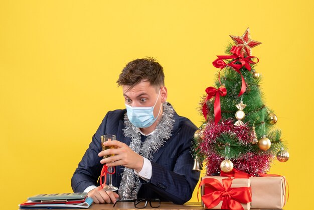 Front view of wide-eyed business man sitting at the table near xmas tree and presents on yellow