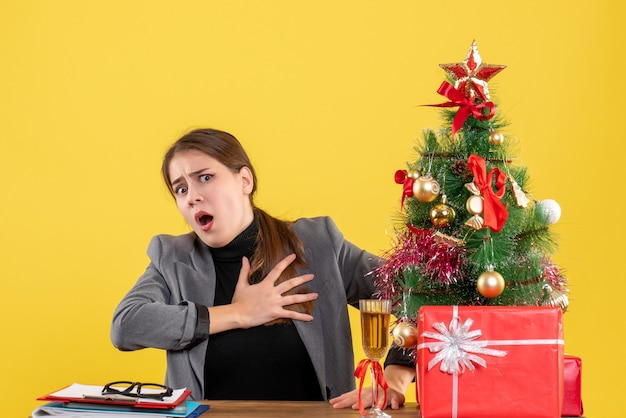 Front view wide-eyed girl sitting at the table holding chest xmas tree and gifts cocktail