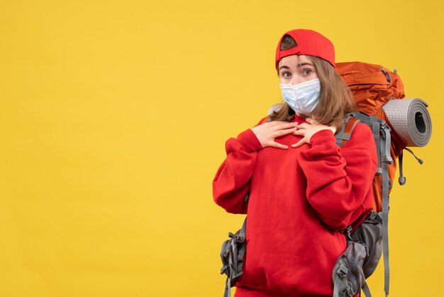 Front view wide-eyed hitchhiker girl with backpack and mask putting hands on her chest