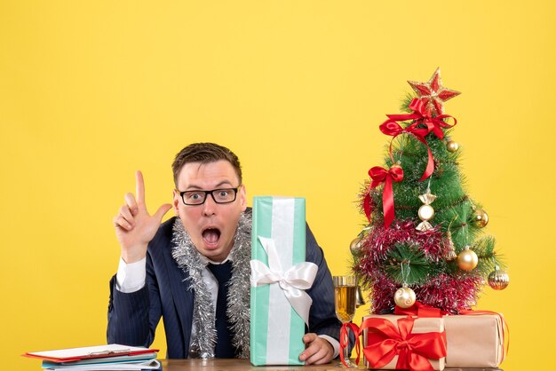 Free photo front view of wide-eyed man pointing at up sitting at the table near xmas tree and presents on yellow