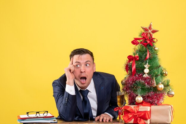 Free photo front view of wide-eyed man sitting at the table near xmas tree and presents on yellow